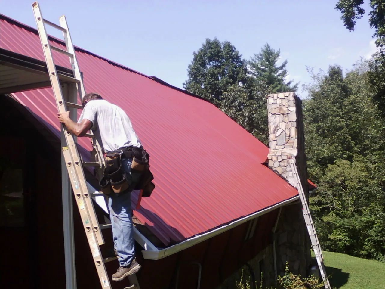 Roofer on a ladder beside a red metal roof and stone chimney — Soffit & Fascia Repair by Roofing Brothers in Harveys Lake